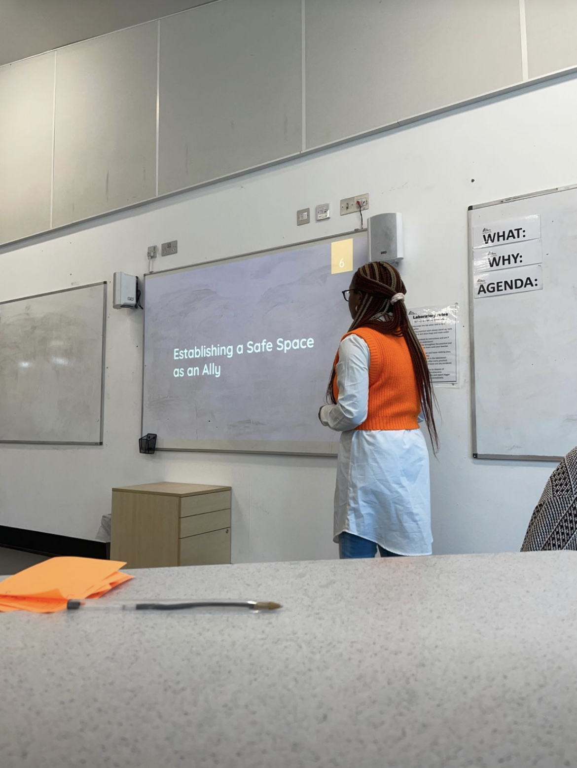 Caring Haven Policy Advisor Mikella is teaching a class on allyship and safe spaces. She is in front of a class, wearing a blue shirt, with orange sweater, and braided hair.